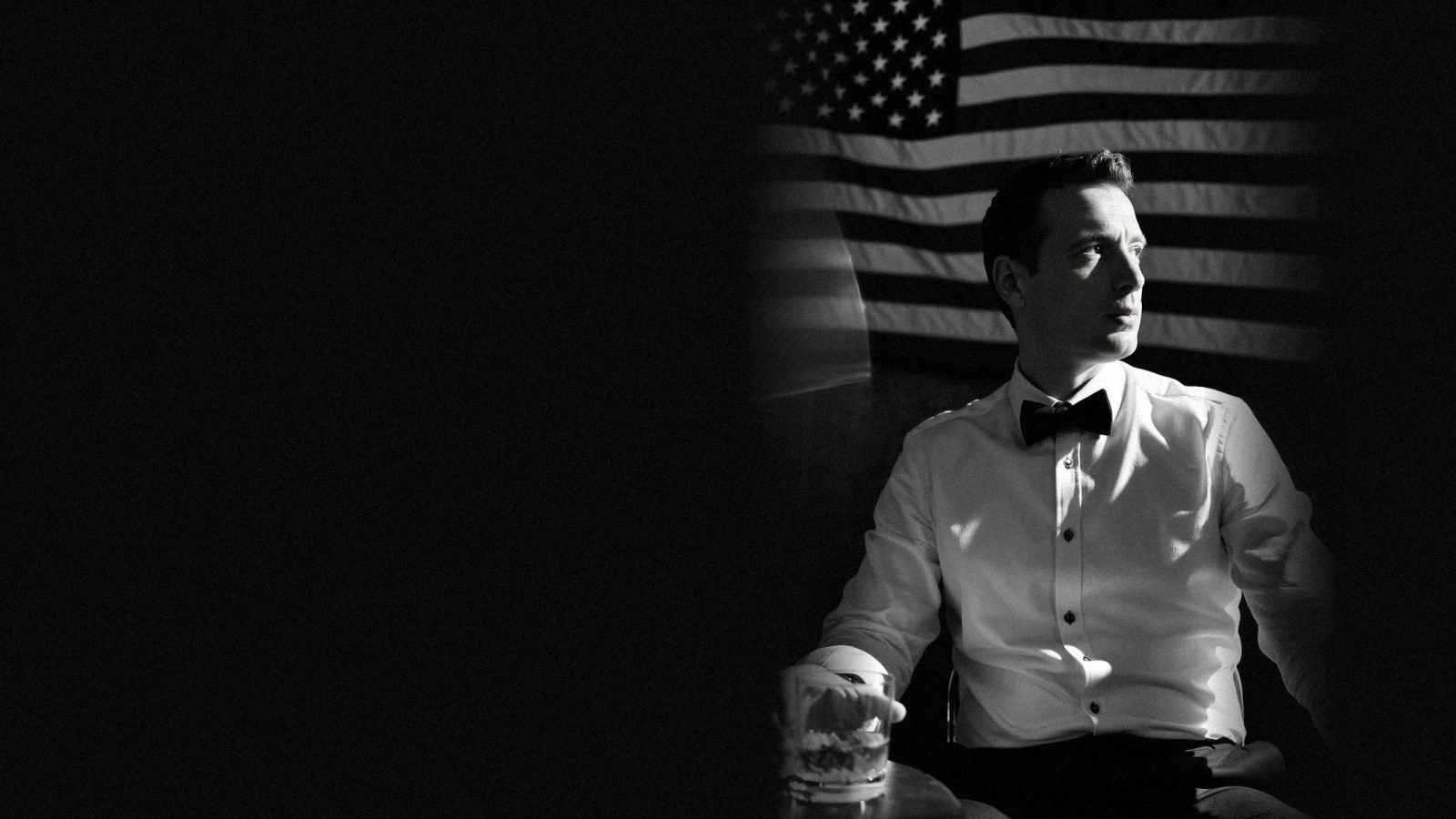 Mikhail in black tie, seated by a window with a flag behind him