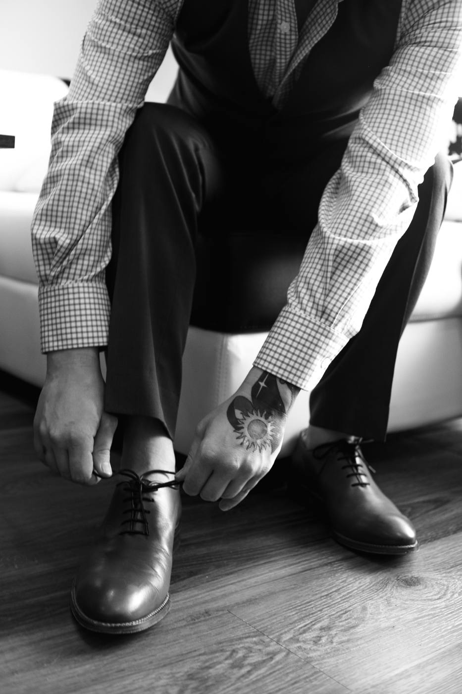Black and white detail of hands lacing a shoe, sun tattoo visible